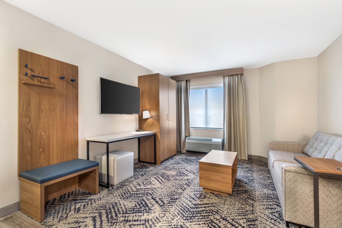 A hotel room with a wooden desk, a bench, a sofa, a TV mounted on the wall, and a large window with curtains.