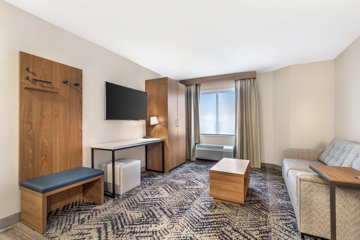 Quarto de hotel com escrivaninha, um banco, sofá, televisão e janela com cortinas.