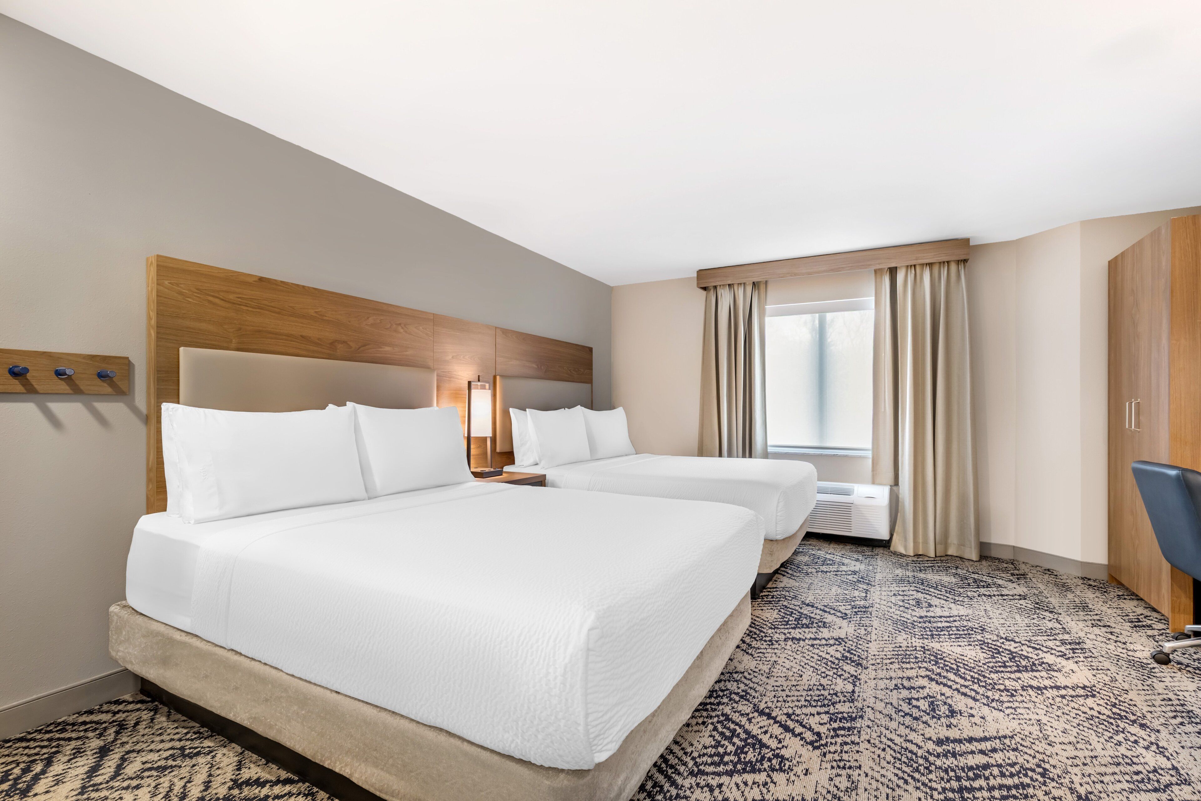 A hotel room with two beds, a wooden headboard, a large window with curtains, a patterned carpet, and a desk with a chair.
