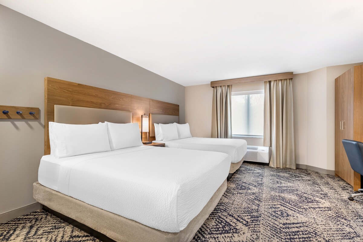 Quarto de hotel com duas camas, cabeceira, mesa de cabeceira, escrivaninha, cadeira, janela com cortinas e tapete estampado.