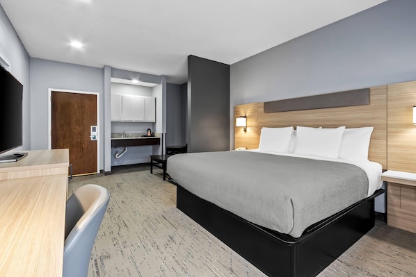 A modern hotel room with a large bed, a desk, a TV, a bathroom, and a kitchenette.