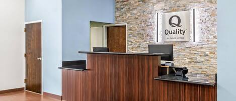 A reception desk with a stone wall backdrop, a sign reading 'Quality by Choice Hotels,' and a wooden door to the left.