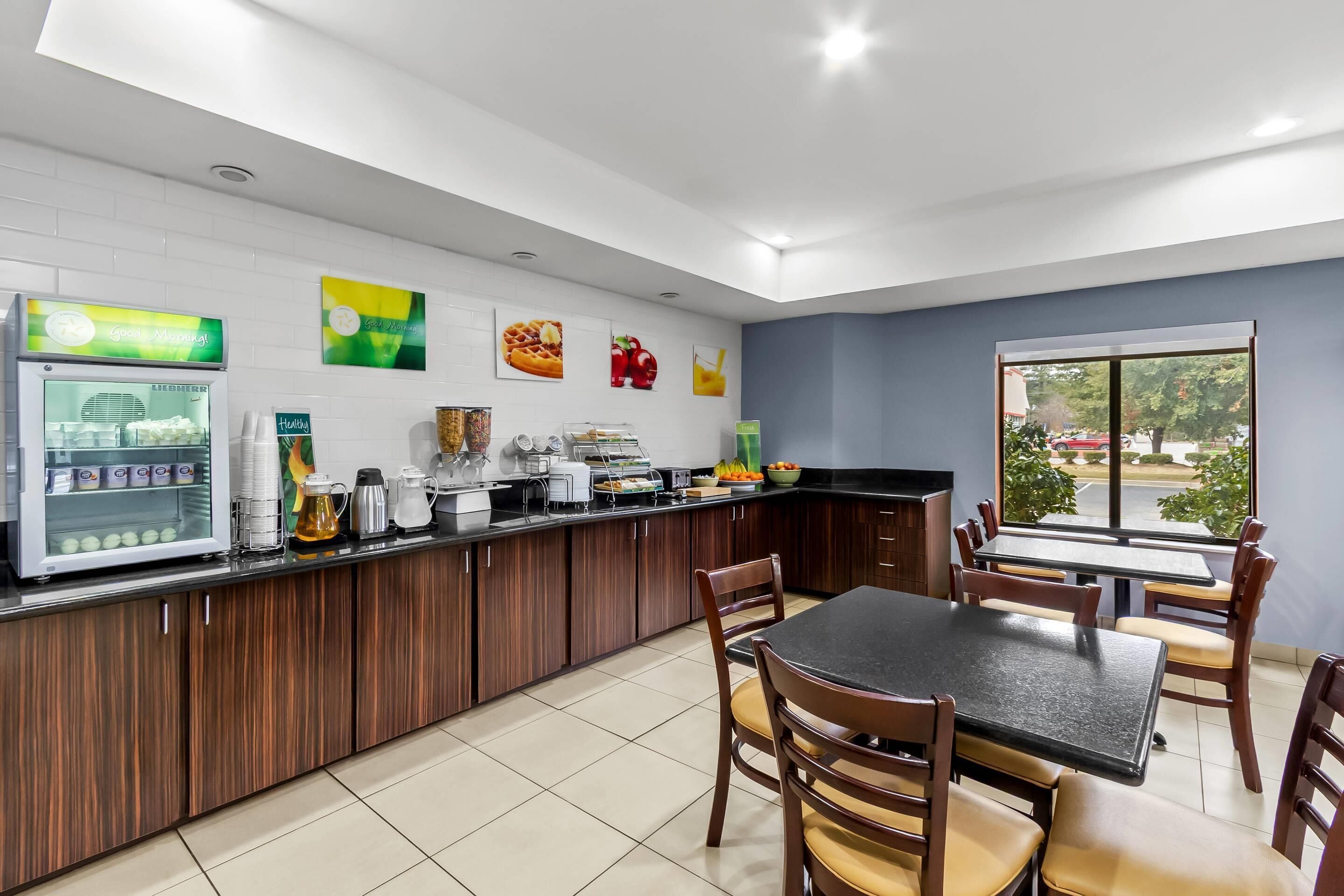 Cozinha moderna com geladeira, cafeteira e balcão com diversos objetos. Há uma mesa de jantar com cadeiras e uma janela com vista para o exterior.
