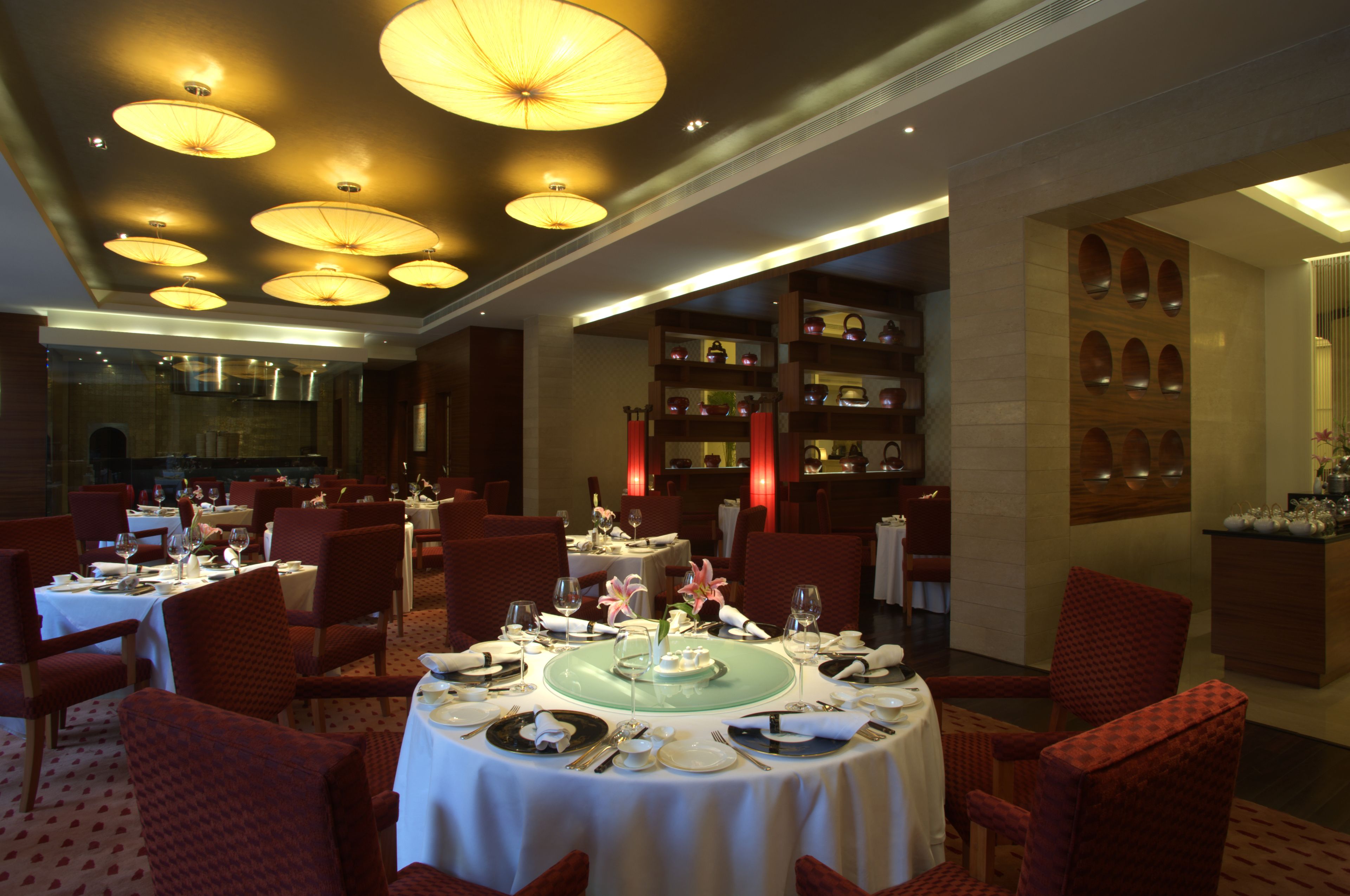 A dining area with round tables set for a meal, red upholstered chairs, and a wall with shelves displaying teapots.