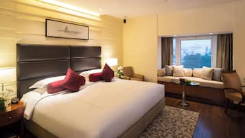 A hotel room with a large bed, a sofa, a small table with fruit, and a window with a city view.
