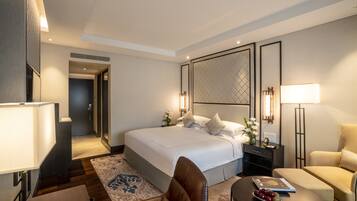 Une chambre d’hôtel moderne avec un grand lit, un coin salon comprenant un canapé et un fauteuil, une petite table avec un livre et des fruits, et un panneau décoratif au mur.