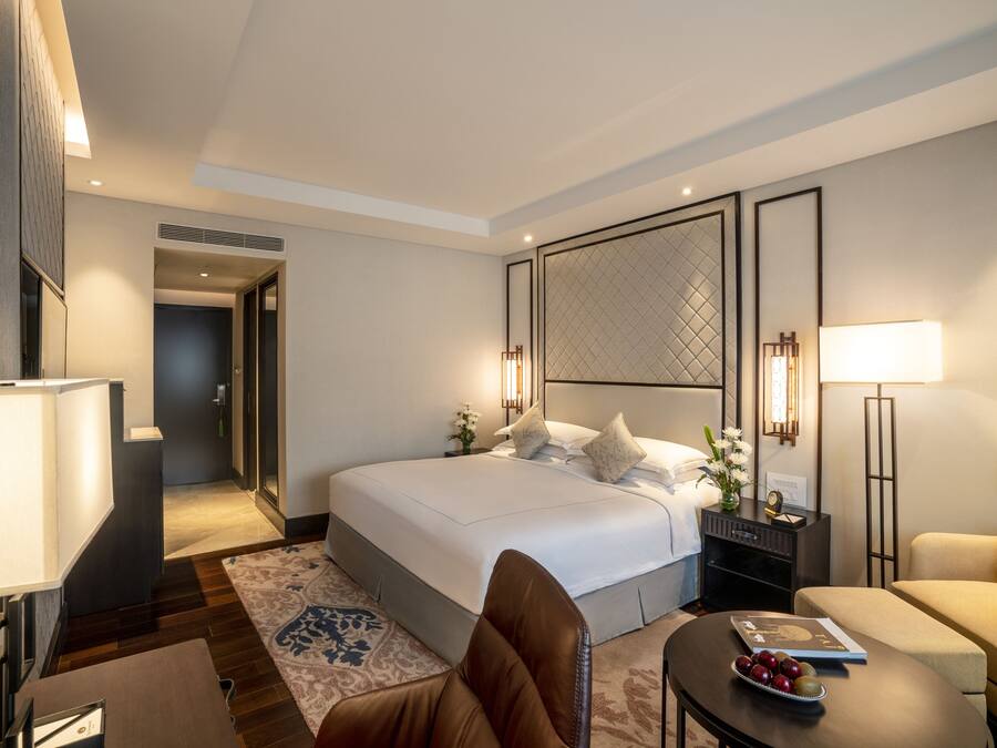 Une chambre d’hôtel moderne avec un grand lit, un coin salon comprenant un canapé et un fauteuil, une petite table avec un livre et des fruits, et un panneau décoratif au mur.