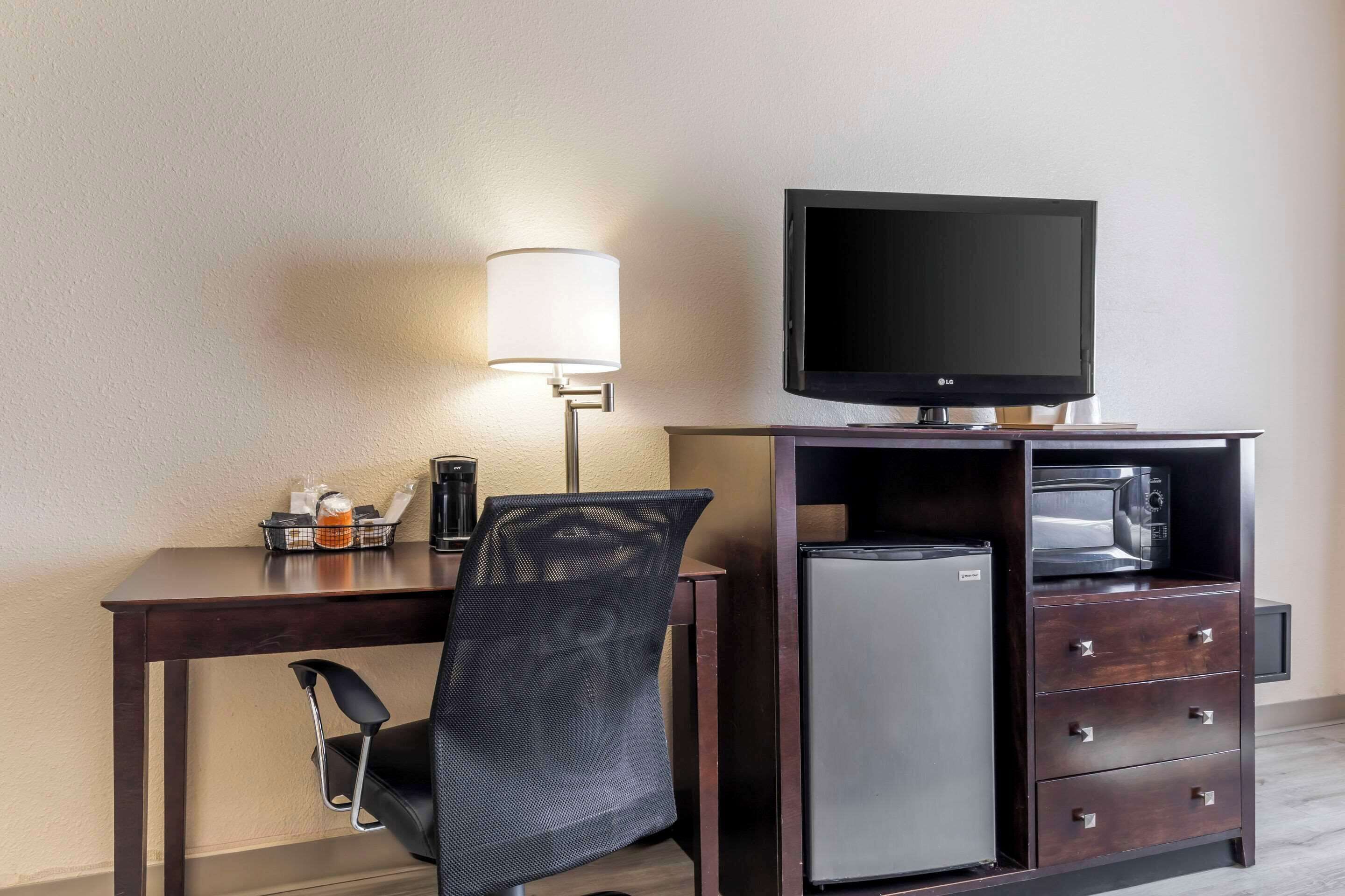 Habitación de hotel con un escritorio de madera oscura, una silla de oficina de malla negra, un televisor de pantalla plana, un microondas, un refrigerador compacto y una lámpara.
