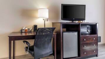 Habitación de hotel con un escritorio de madera oscura, una silla de oficina de malla negra, un televisor de pantalla plana, un microondas, un refrigerador compacto y una lámpara.