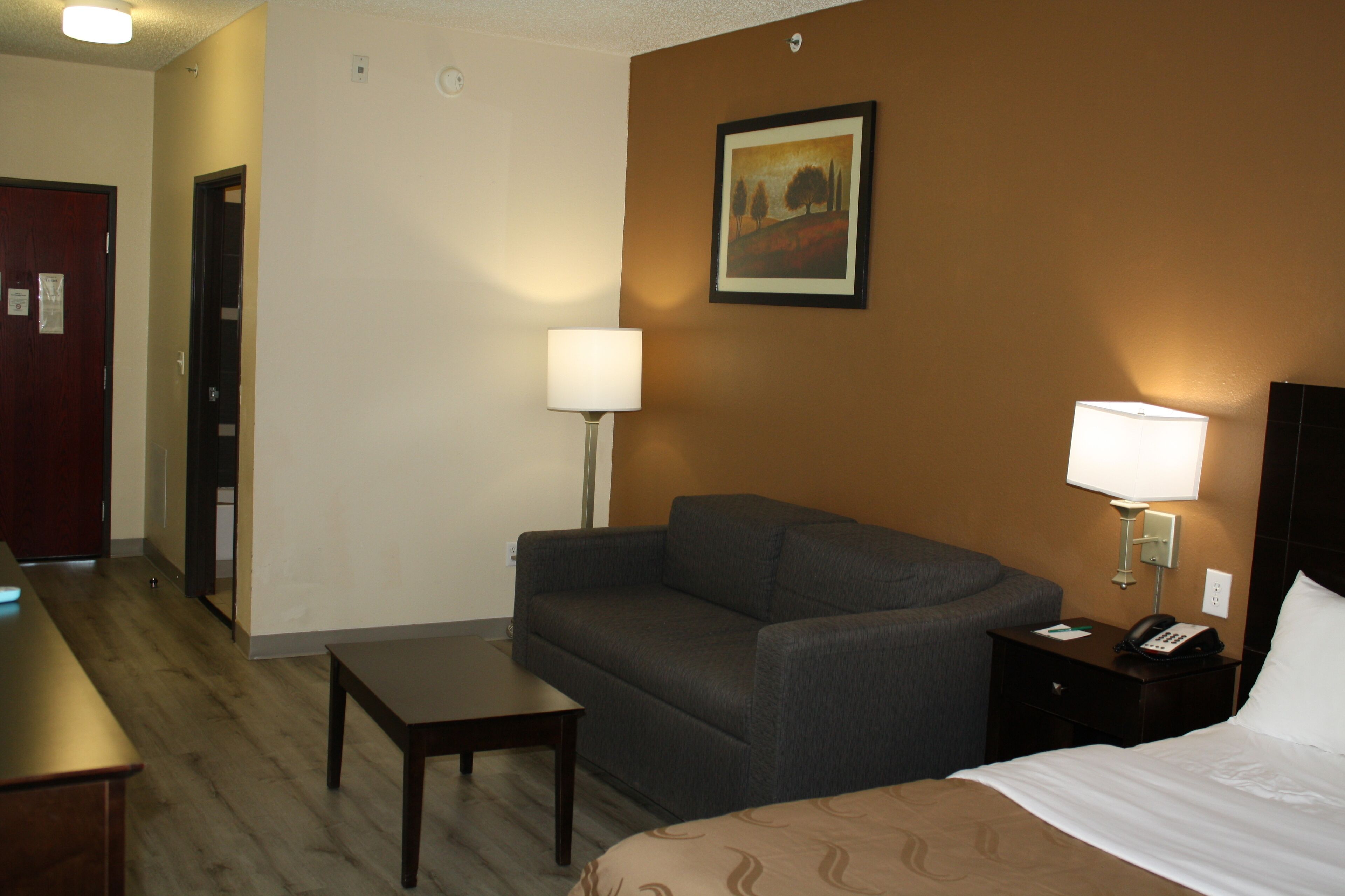 A hotel room with a bed, a sofa, a small table, a nightstand with a lamp, and a painting on the wall.