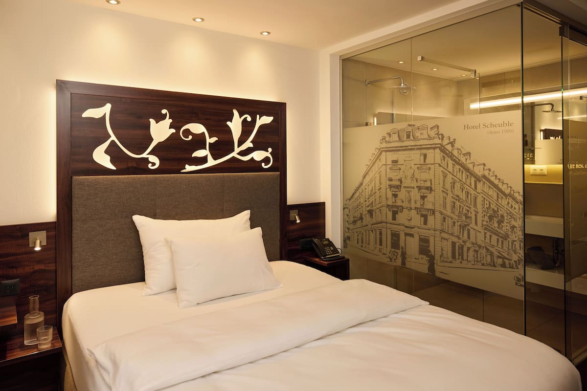 A hotel room with a large bed, a headboard with decorative elements, a nightstand with a phone, and a glass wall with a cityscape mural.