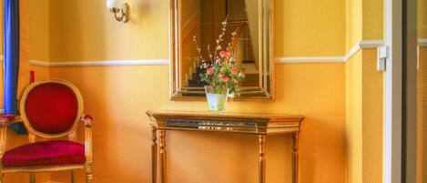 Ein Zimmer mit einem rot gepolsterten Sessel, einem Holzkonsolentisch mit Blumen in einer Vase und einem Spiegel an der Wand.