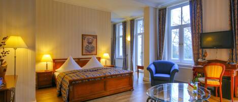 A hotel room with a wooden bed, two bedside tables with lamps, a glass coffee table, a blue armchair, and a television on the wall.