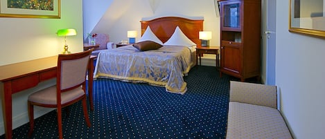 A hotel room with a bed, a desk, a chair, a nightstand, a lamp, and a cabinet.