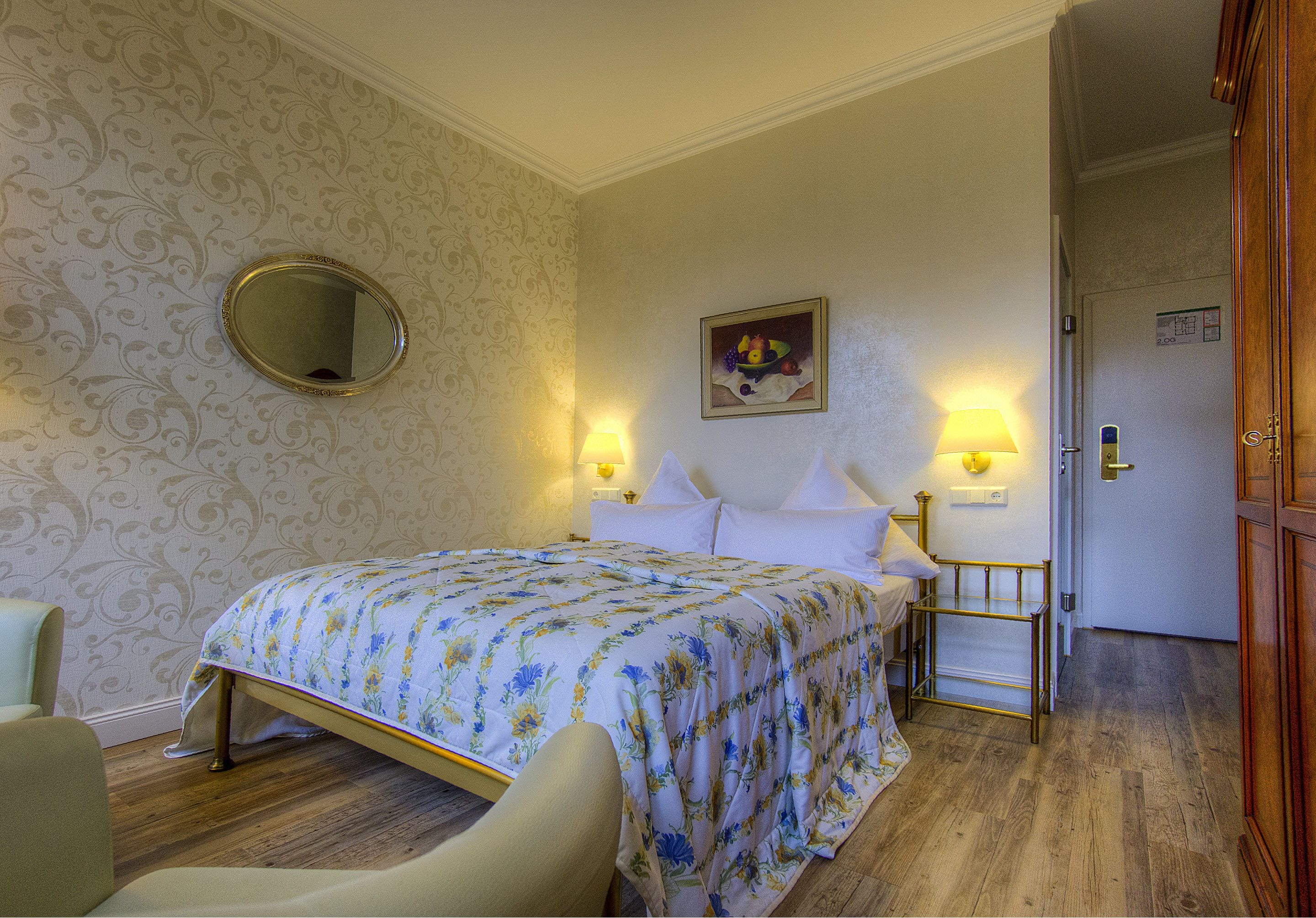 A hotel room with a bed, a round mirror, a painting, two bedside lamps, and a door.