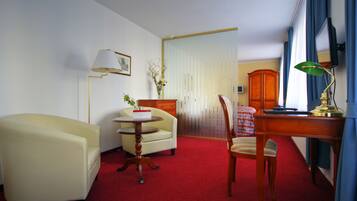 Quarto de hotel com carpete vermelho, poltrona amarela, mesa redonda, escrivaninha de madeira, cadeira, armário e uma janela grande com cortinas.