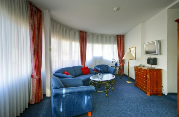 Ein Hotelzimmer mit geschwungener Anordnung, ausgestattet mit einer blauen Sofagarnitur, einem runden Couchtisch, einer Holzkommode und einem an der Wand befestigten Fernseher.
