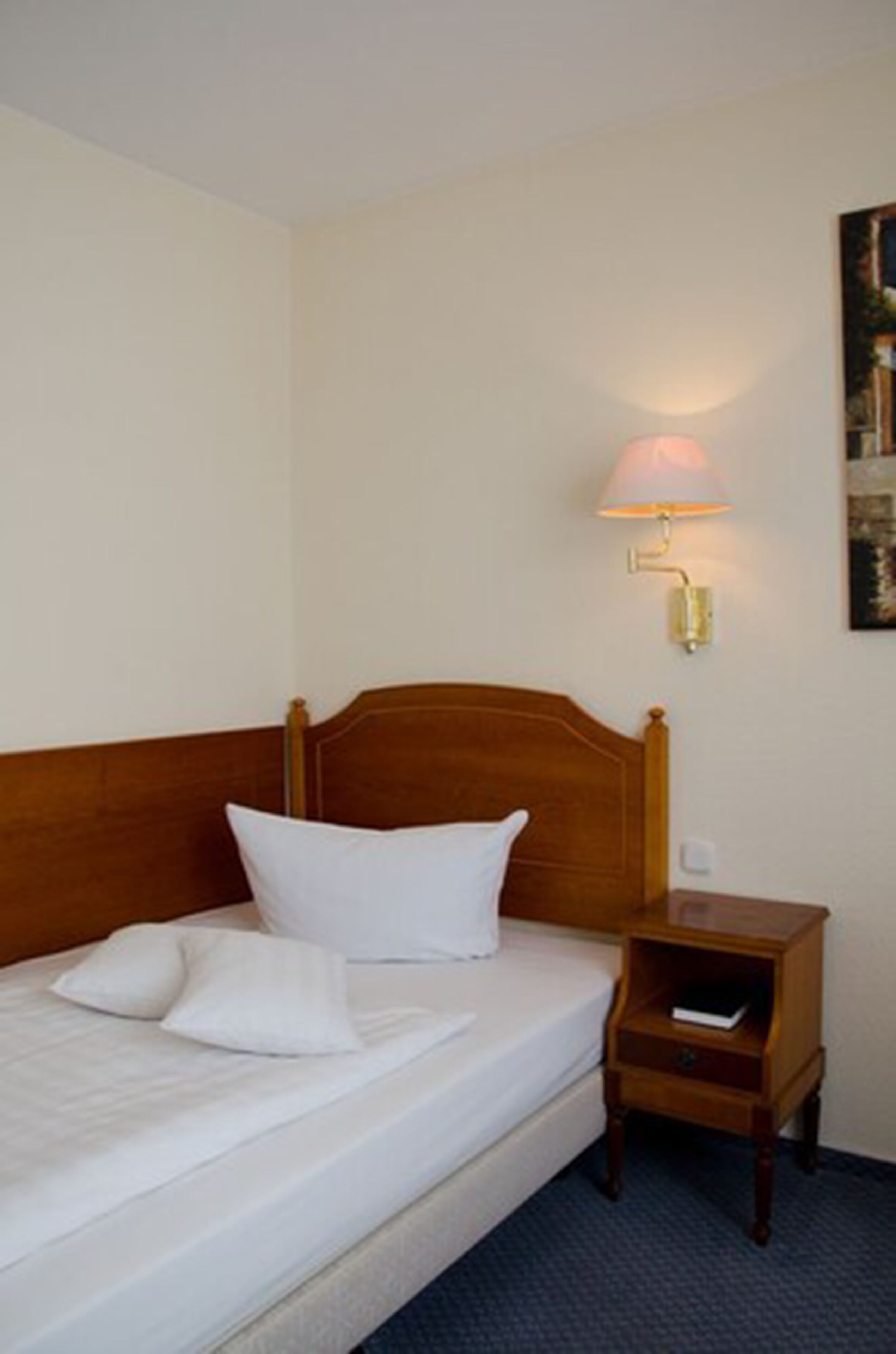 Una cama bien hecha con sábanas y almohadas blancas, un cabecero de madera, una mesita de noche con una lámpara y un cuadro en la pared.