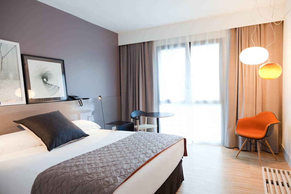 A hotel room with a large bed, a desk, a chair, and a window with curtains.