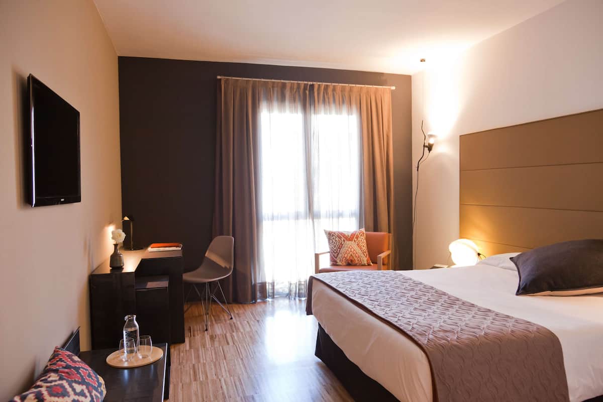 A hotel room with a bed, a desk, a chair, a TV, and a window with curtains.
