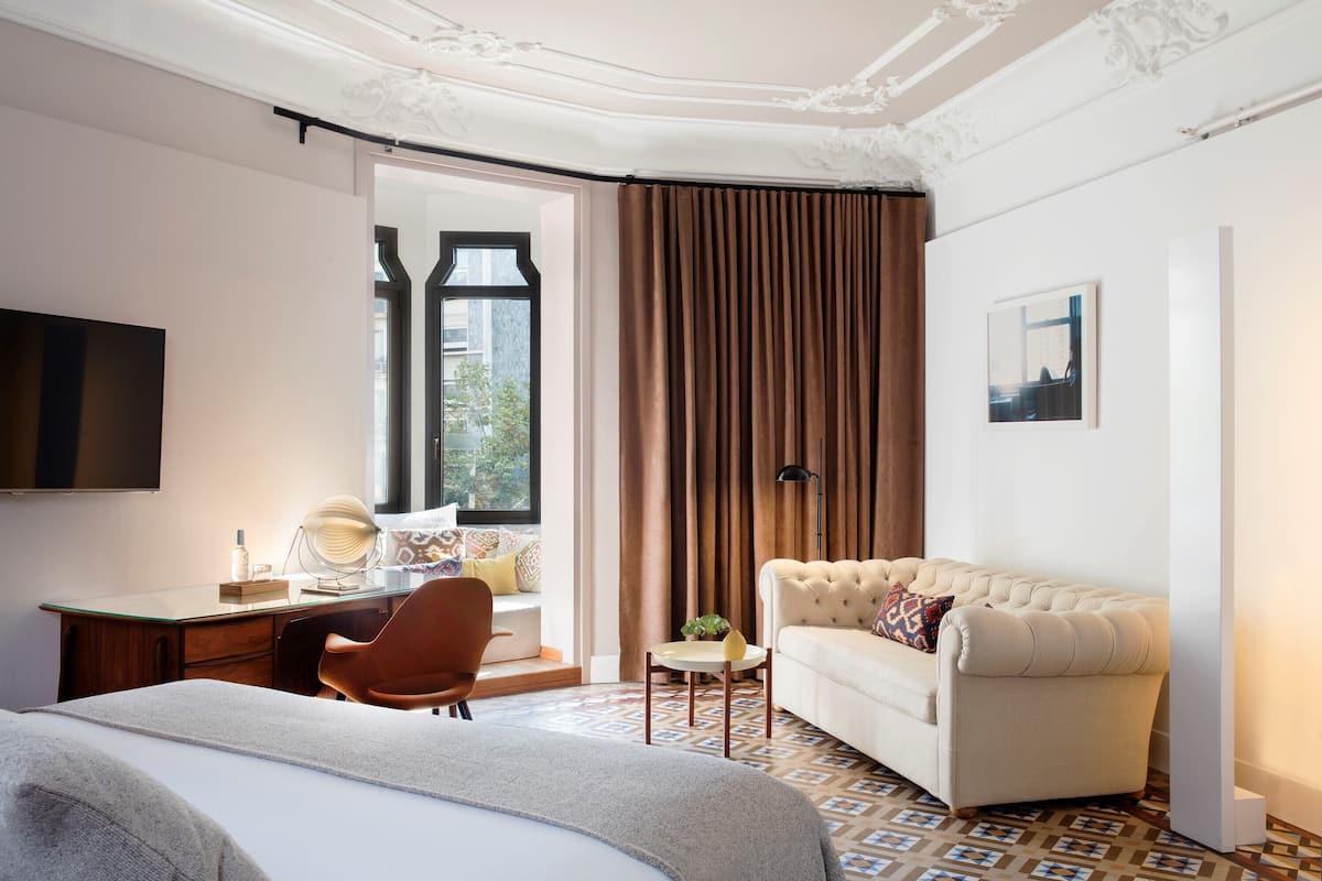 A hotel room with a bed, a desk, a chair, a sofa, a television, and a window with curtains.