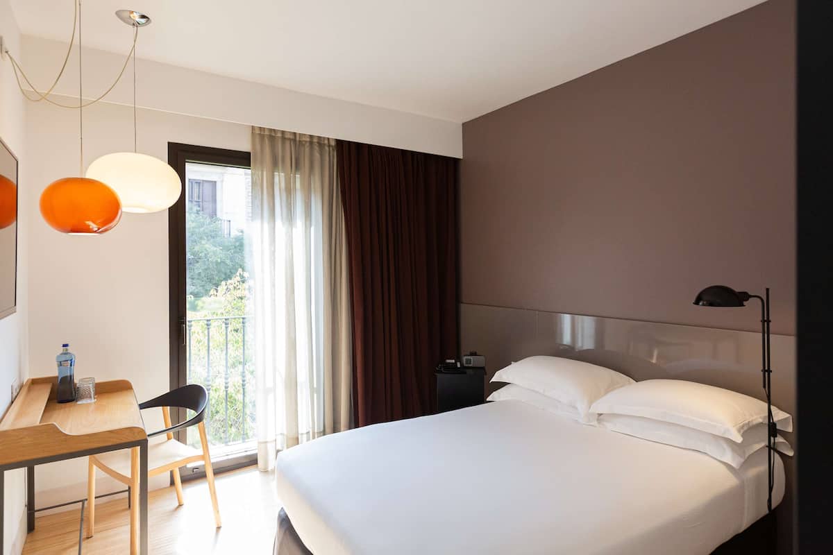A hotel room with a bed, a desk with a chair, a table with a bottle and glass, a nightstand with a lamp, and a view of greenery through the window.