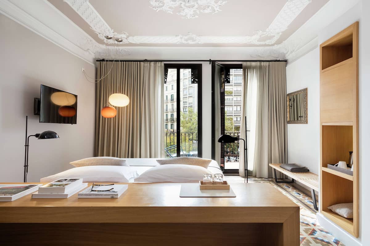 A modern hotel room with a large bed, a desk with books, a TV, and a view of the city through large windows.