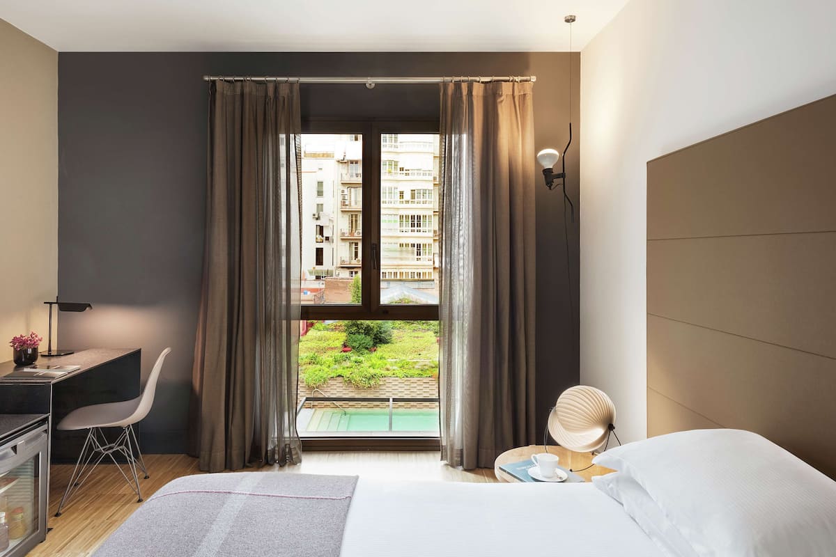 A hotel room with a bed, a desk, a chair, a lamp, and a view of a pool and buildings.