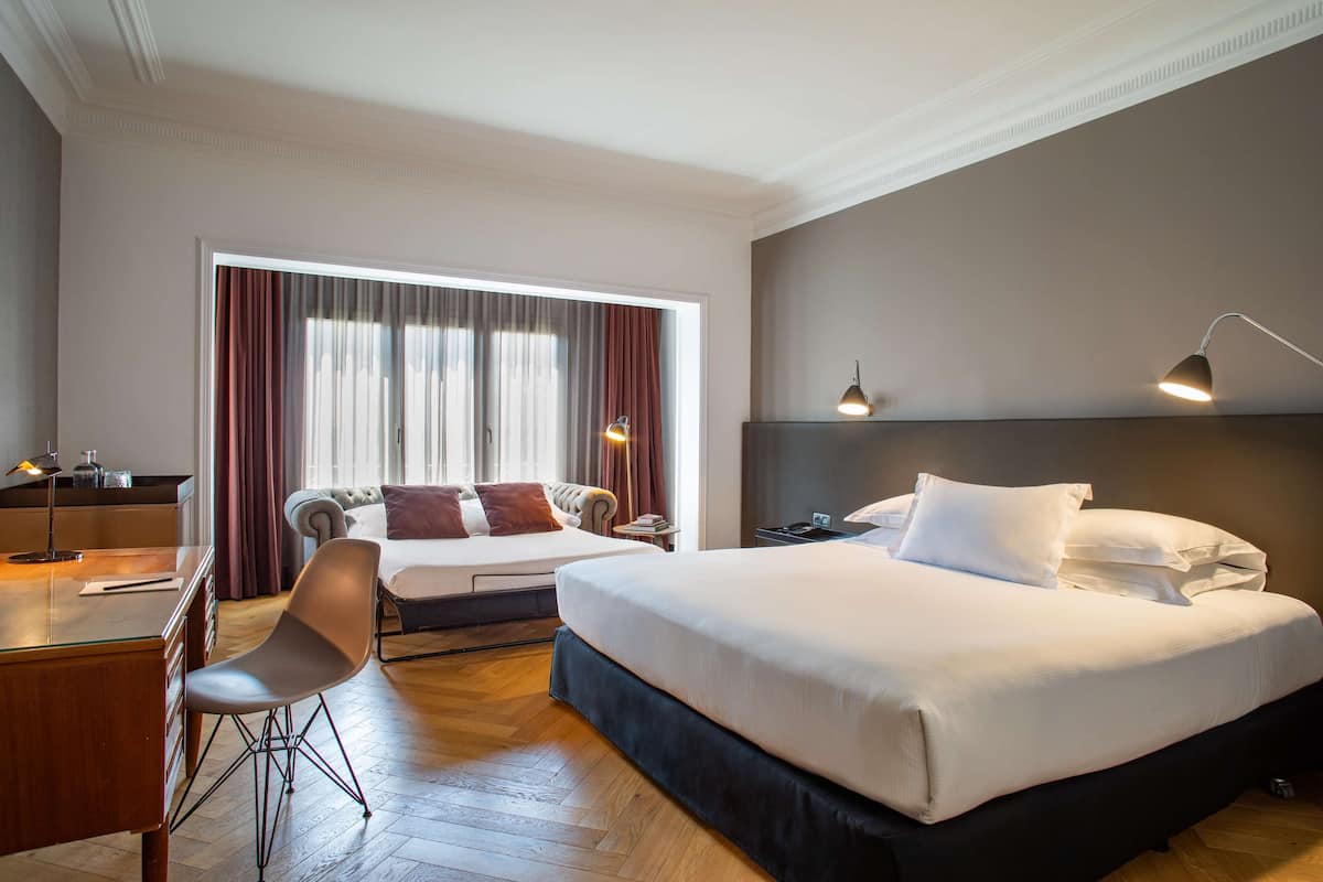 A hotel room with a large bed, a desk with a chair, a lamp, and a view of the city through the window.