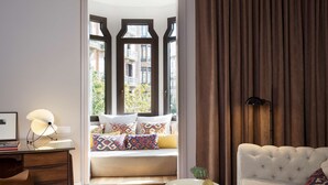 A modern hotel room with a sofa bed, a desk with a lamp, and a view of the city through large windows. - Alexandra Barcelona Hotel, Curio Collection by Hilton (Barcelona)