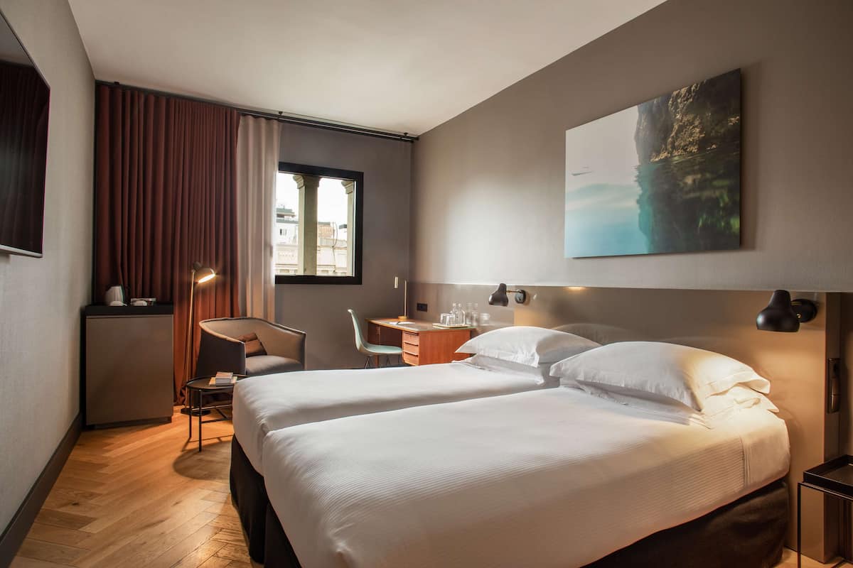 A hotel room with two beds, a desk, a chair, and a large window.
