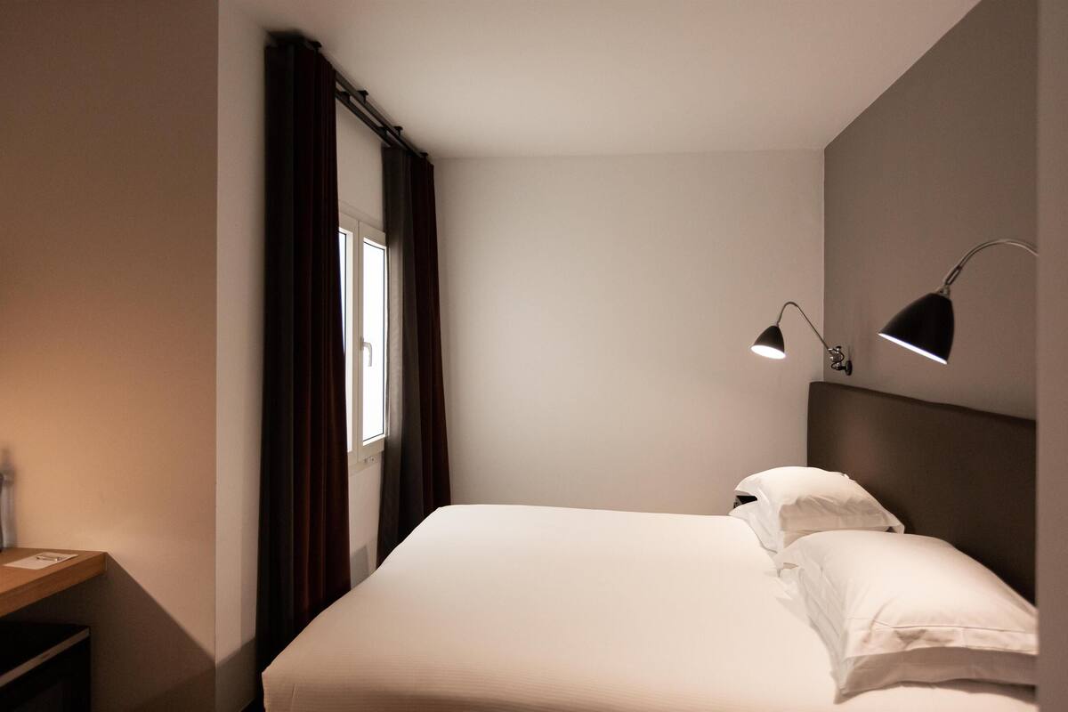 A hotel room with a bed, a nightstand, a desk, and a window with curtains.