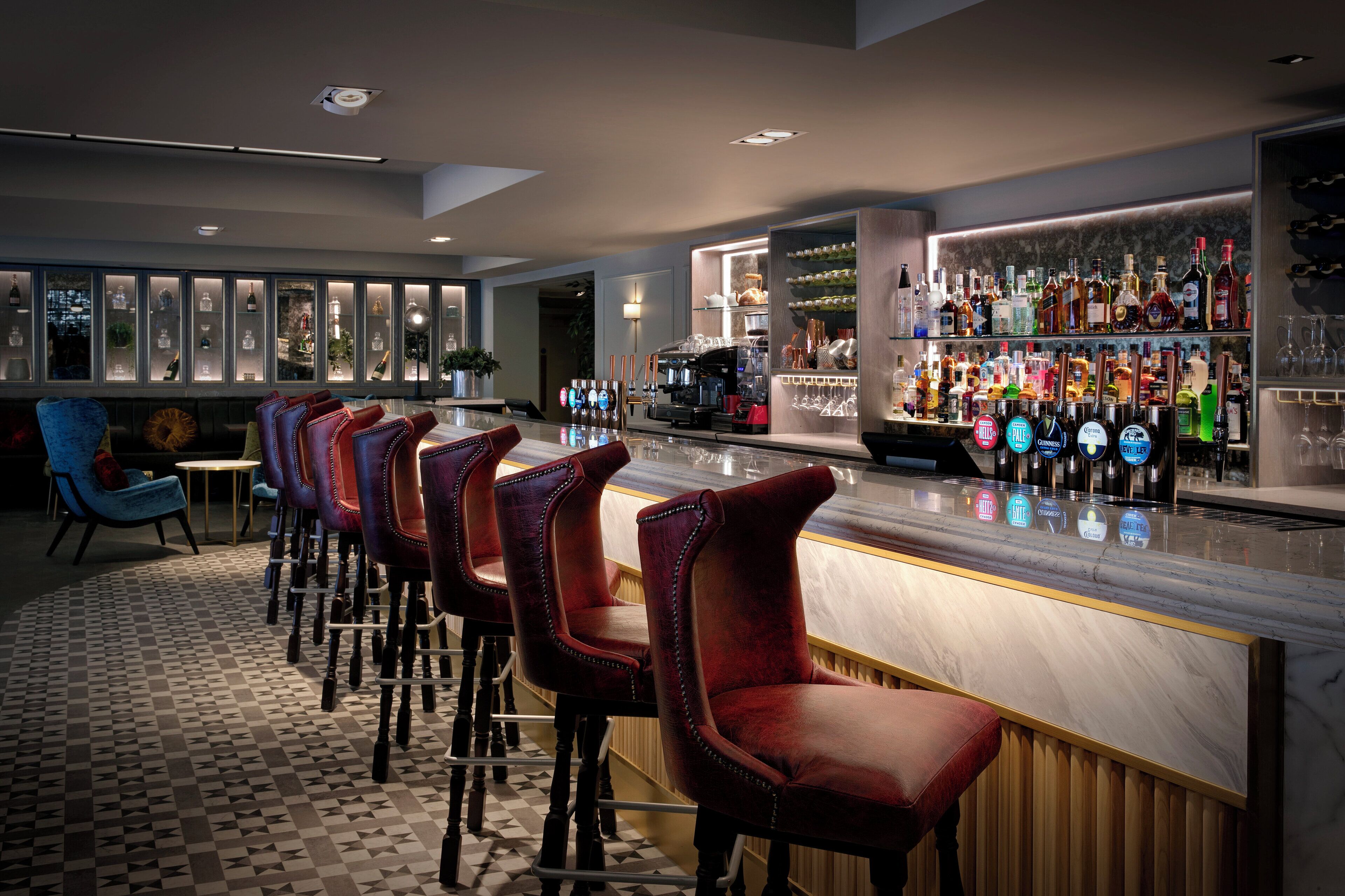 A bar area with a marble countertop, red upholstered bar stools, and a variety of liquor bottles and glasses.