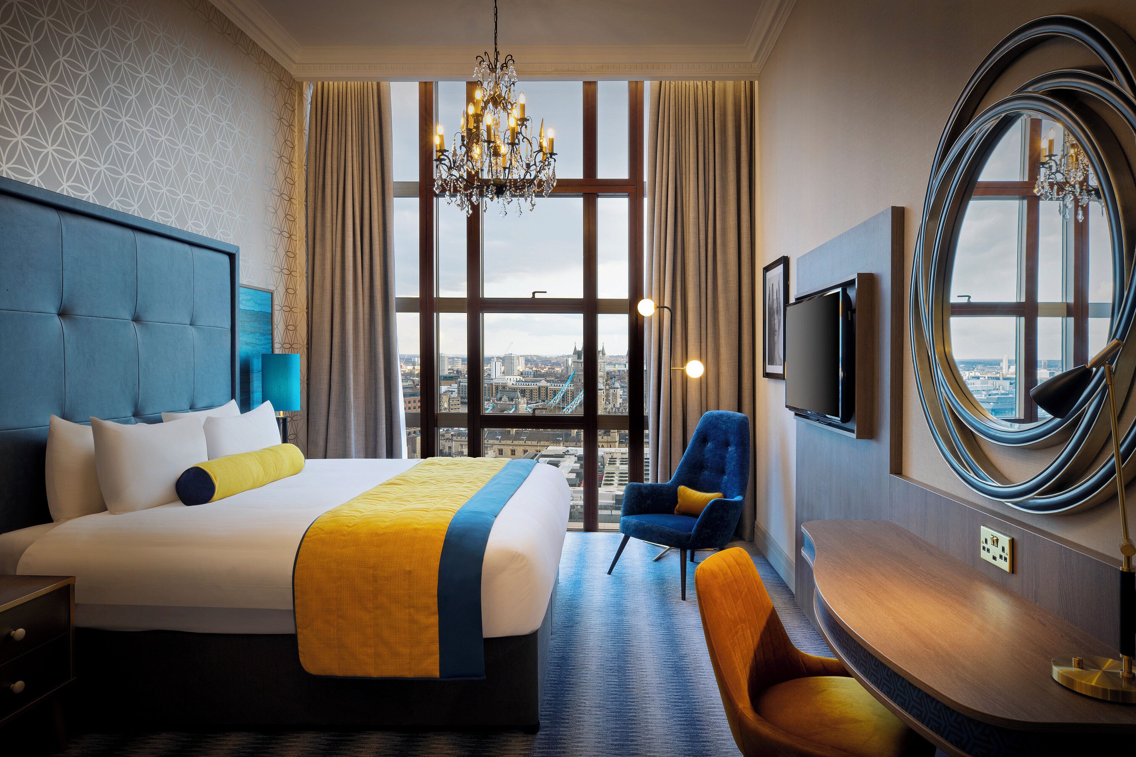 A hotel room with a large bed, a TV, a chair, and a view of the city through large windows.
