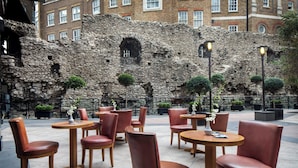 An outdoor seating area with round tables and leather chairs, set against a stone wall with a window and potted plants. - Leonardo Royal Hotel London City - Tower of London (London)