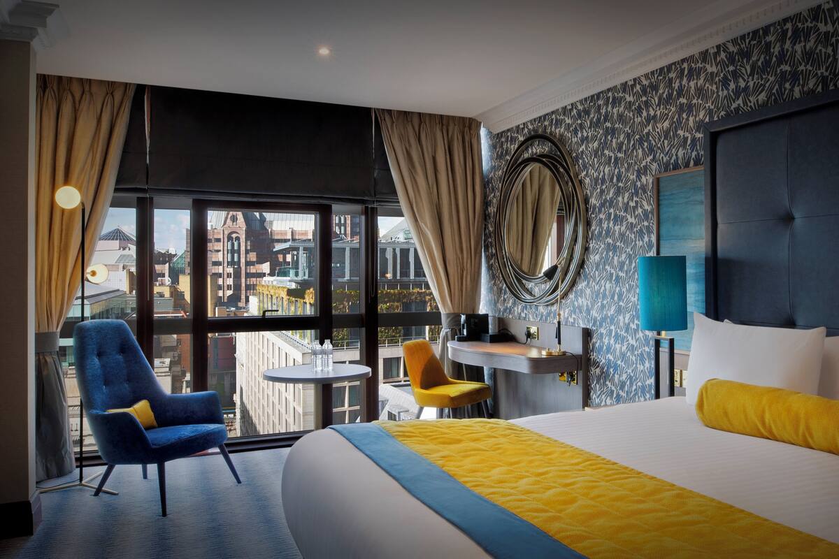 Une chambre d’hôtel avec un grand lit, un bureau, deux chaises, un grand miroir rond et une vue sur le paysage urbain grâce à de grandes fenêtres.