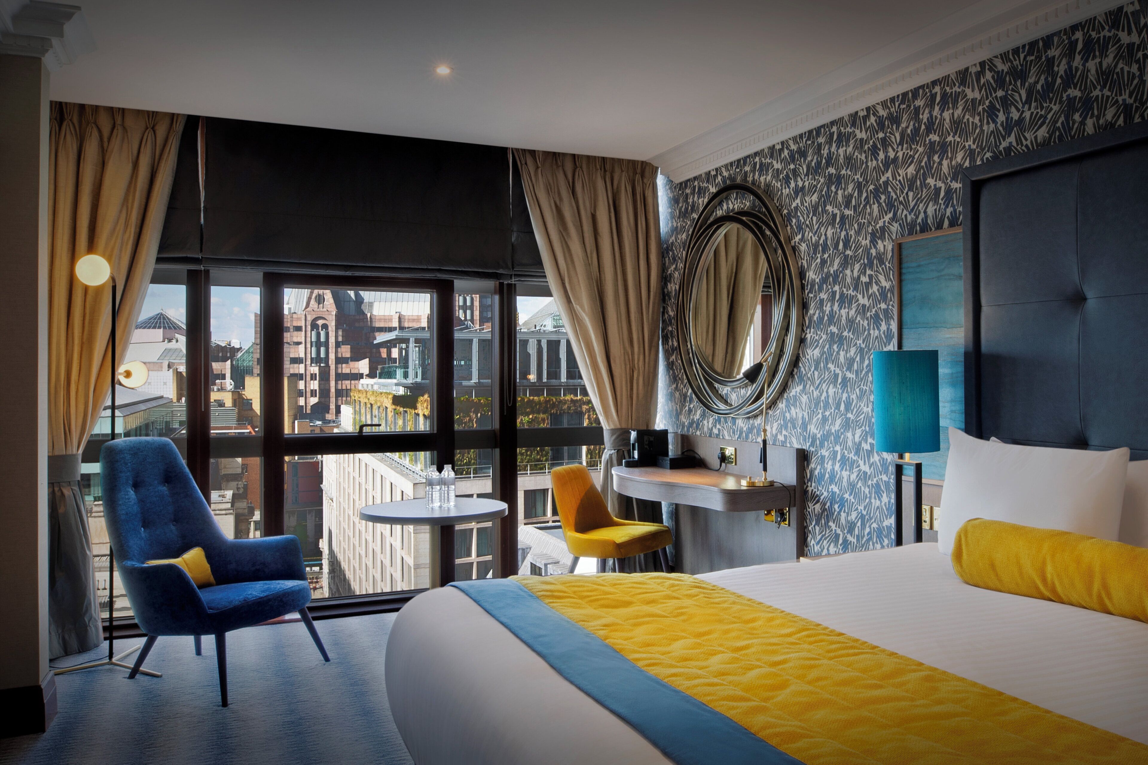 A hotel room with a large bed, a desk, two chairs, a round mirror, and a view of the cityscape through large windows.
