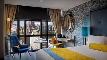 A hotel room with a large bed, a desk, two chairs, a round mirror, and a view of the cityscape through large windows.