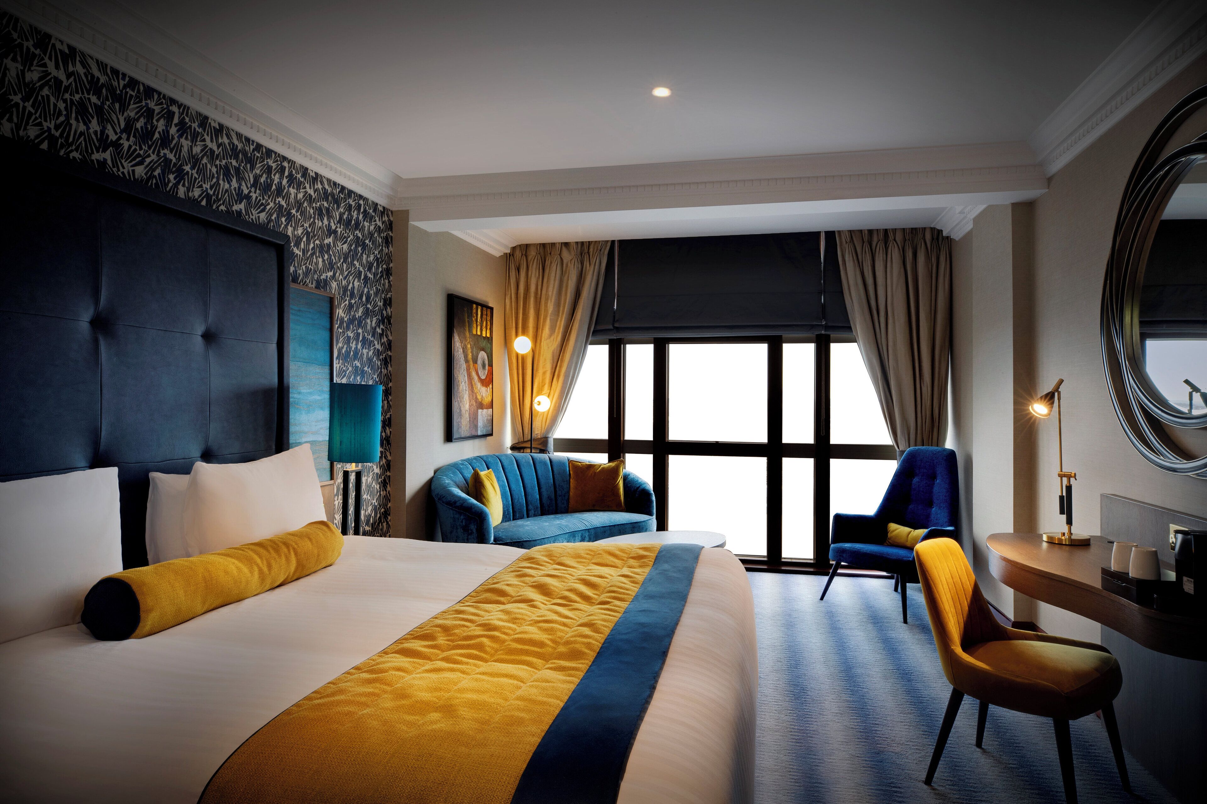 Una habitación de hotel con una cama grande, un escritorio, dos sillas y un ventanal amplio.