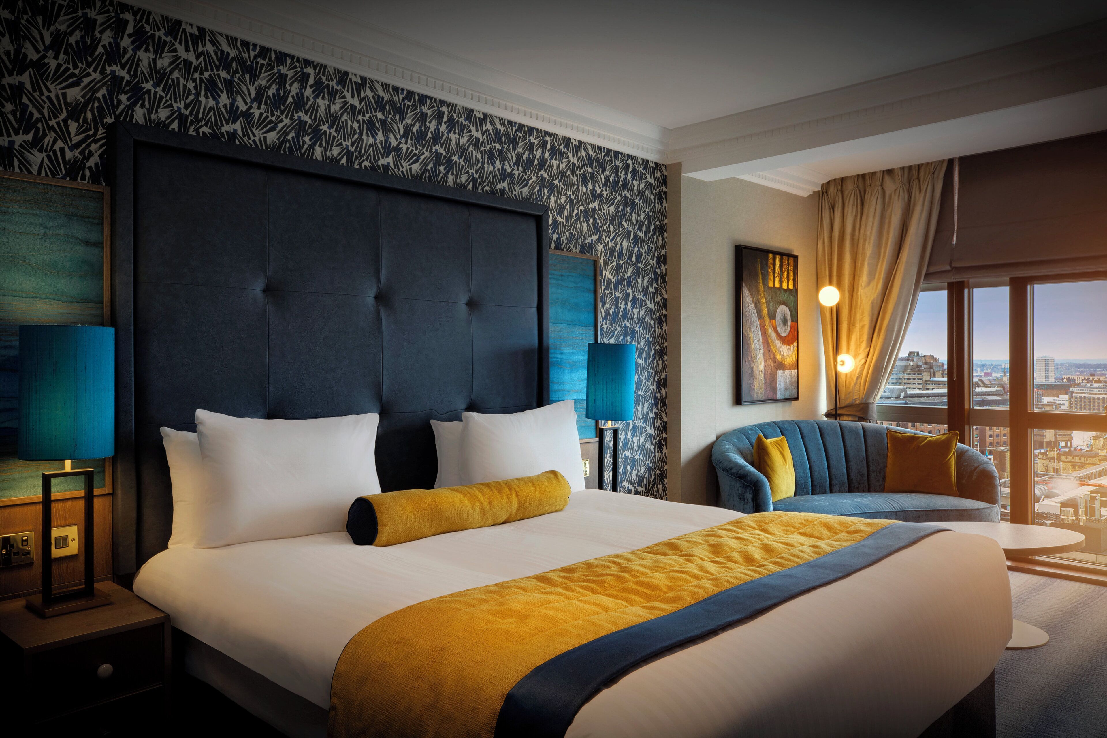 Ein Hotelzimmer mit einem großen Bett, einer Sitzecke, Blick auf die Stadt und einer Wand mit Muster.