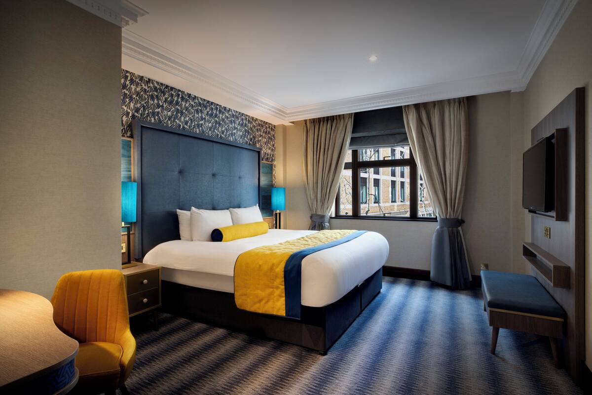 Une chambre d’hôtel avec un grand lit, une table de chevet, une chaise, une télévision et une fenêtre avec des rideaux.