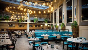 A spacious indoor dining area with multiple seating arrangements, including round tables and a long blue sofa. - Leonardo Royal Hotel London City - Tower of London (London)