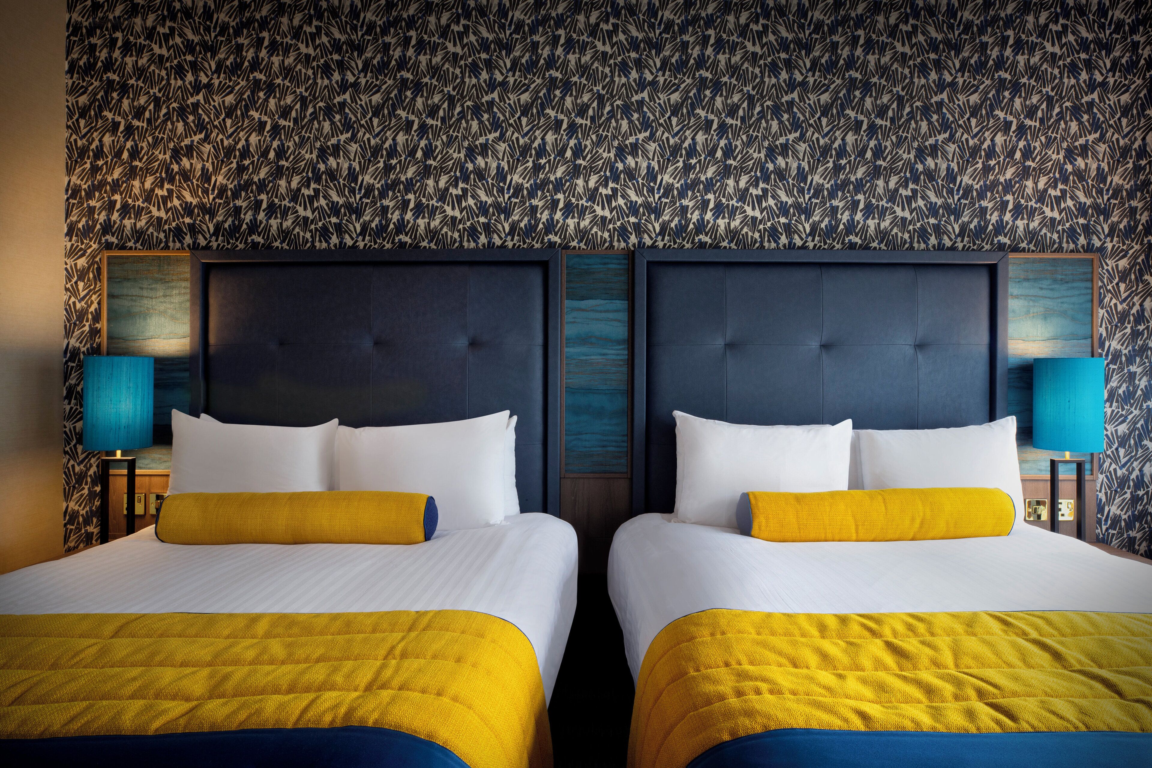 A hotel room with two beds, each with a yellow and white bedspread, and a dark headboard. The room has a patterned wallpaper and blue accent lighting.