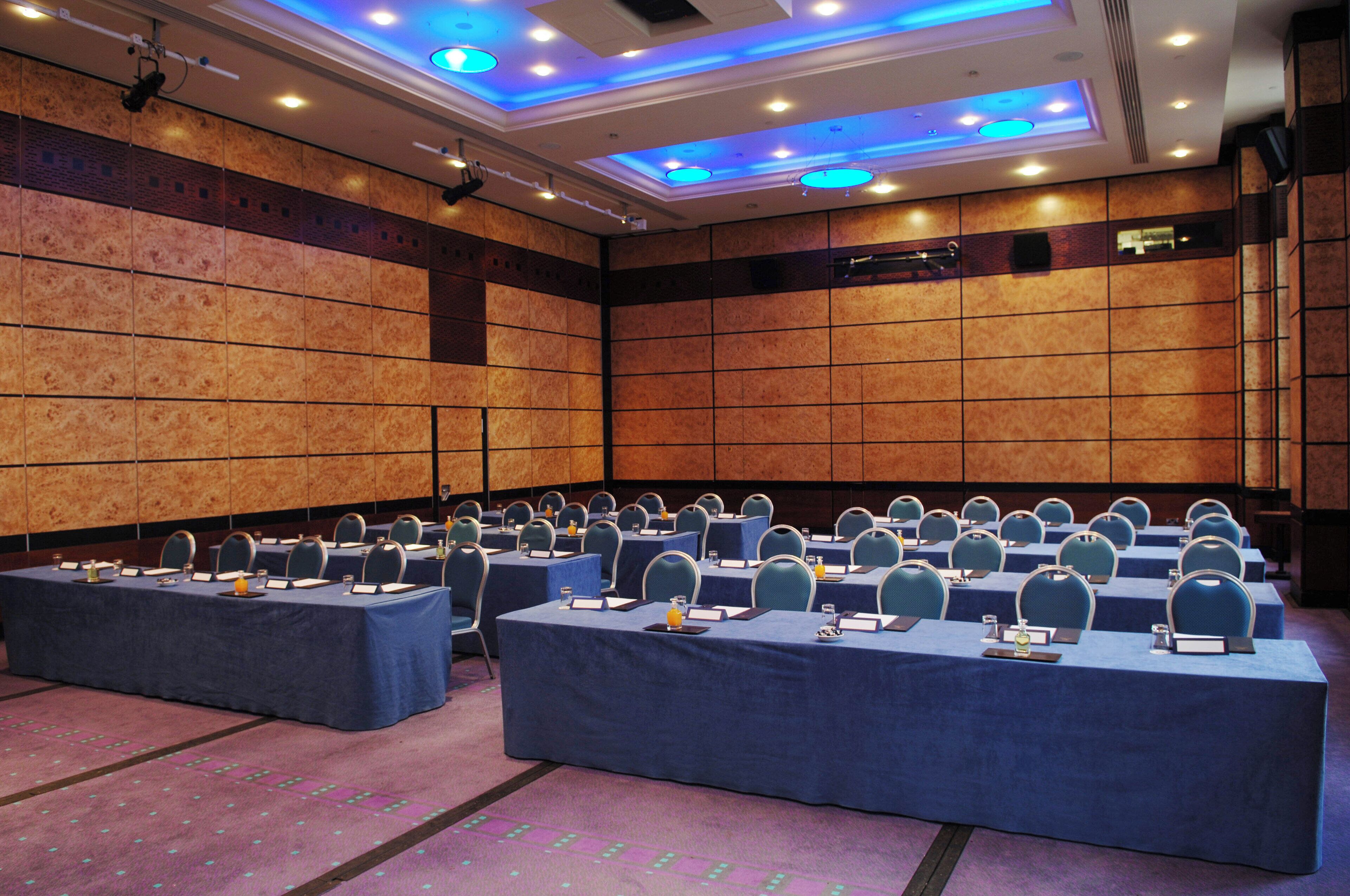 A conference room with multiple tables, chairs, and nameplates, featuring a high ceiling with recessed lighting and a patterned wall.