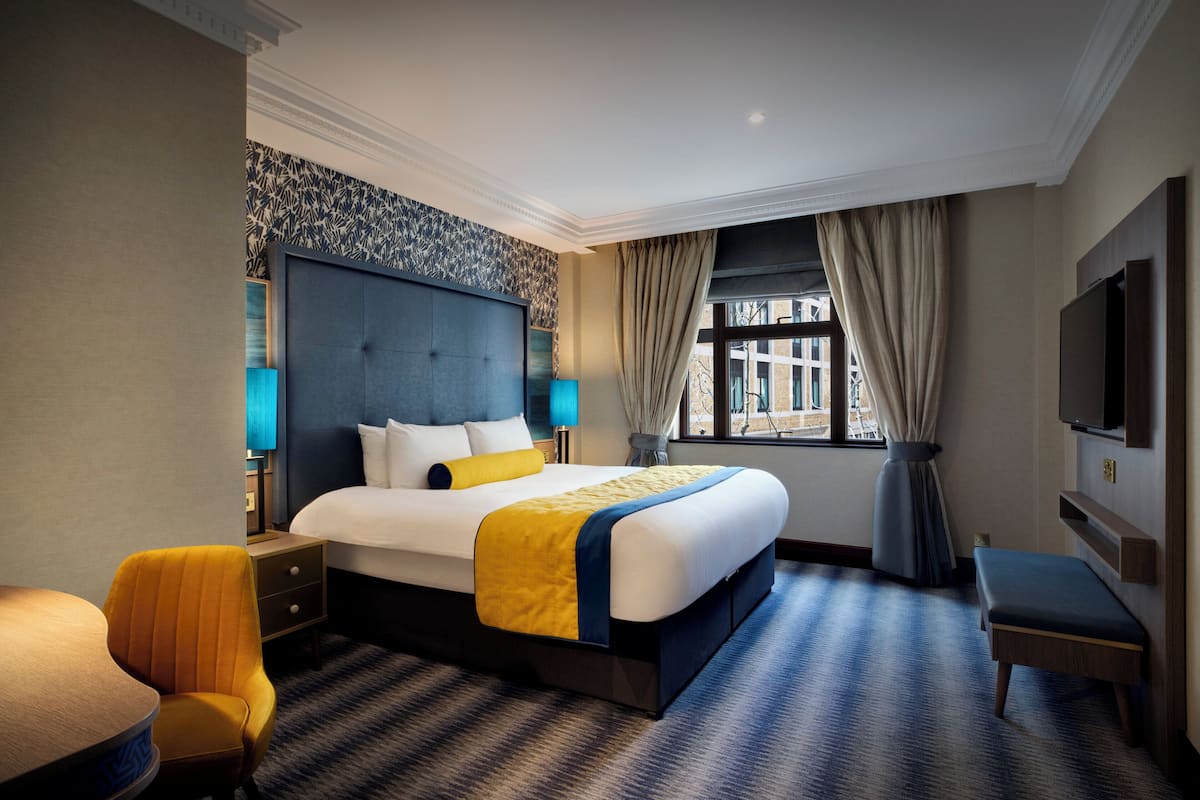 A hotel room with a large bed, a bedside table, a chair, a TV, and a window with curtains.