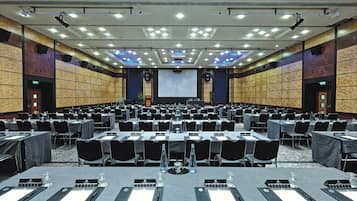 A large conference room with rows of tables and chairs, a large screen at the front, and a stage with a podium.
