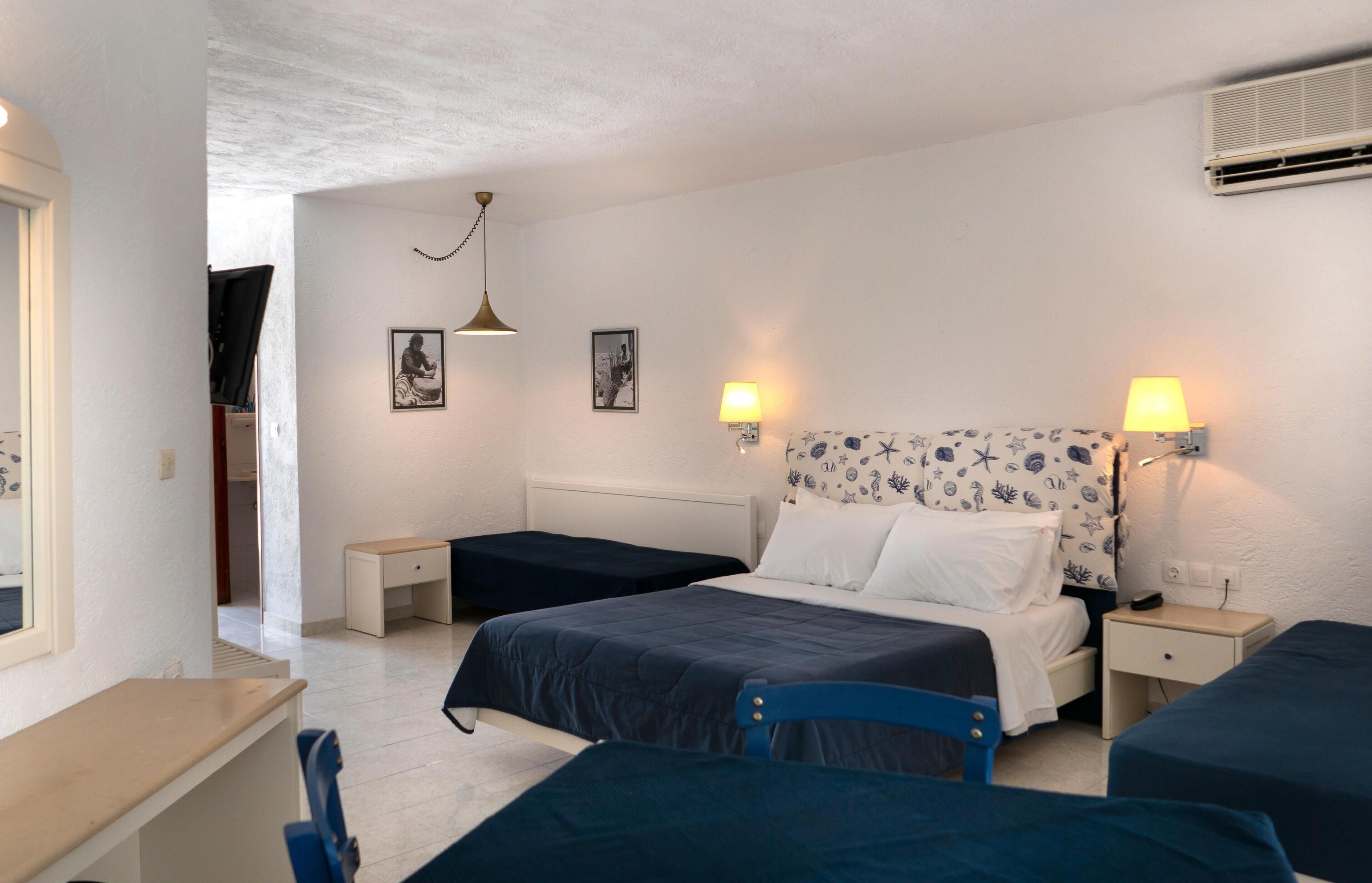 A bedroom with two single beds, a desk, and a chair. There are two bedside tables with lamps, a ceiling light, and framed pictures on the wall.