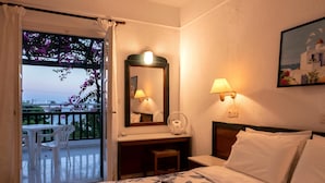 A hotel room with a balcony, a bed with floral bedding, a desk with a mirror, and a nightstand with a lamp. - Villea Village by GHH (Sitia)