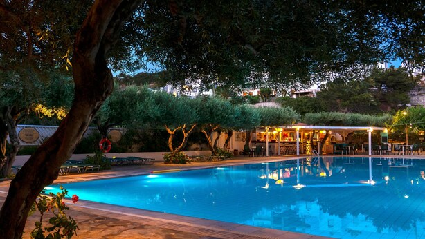 A swimming pool with lounge chairs and a bar area, surrounded by trees and a building in the background. - Villea Village by GHH (Sitia)