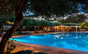 A swimming pool with lounge chairs and a bar area, surrounded by trees and a building in the background. - Villea Village by GHH (Sitia)